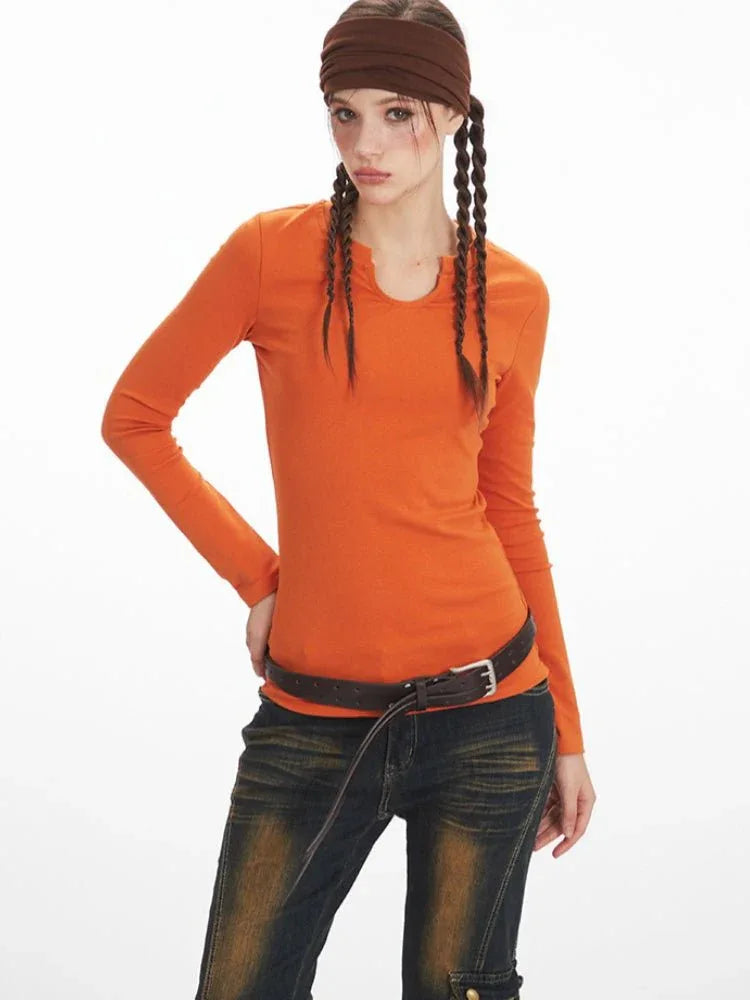 Close-up front view of a woman wearing an orange long-sleeve notch neck top and dark wash distressed jeans, accessorized with a brown belt.