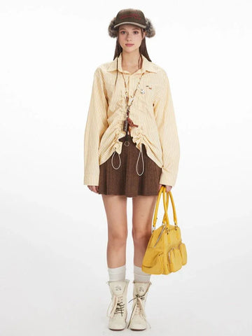 Woman wearing a yellow and white striped long-sleeved collared shirt with ruched drawstring detail, a brown knit skirt, white socks, and white boots, holding a yellow handbag.