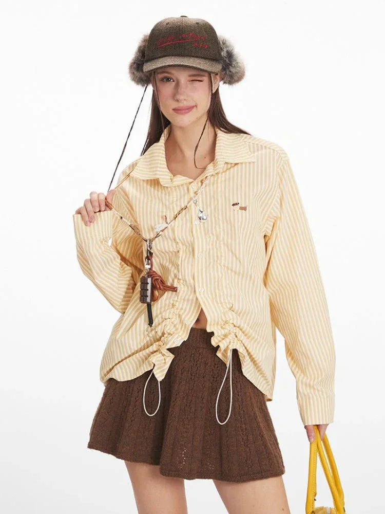 Woman wearing a yellow and white striped long-sleeved collared shirt with ruched drawstring detail and a brown knit skirt, winking and holding the drawstring.