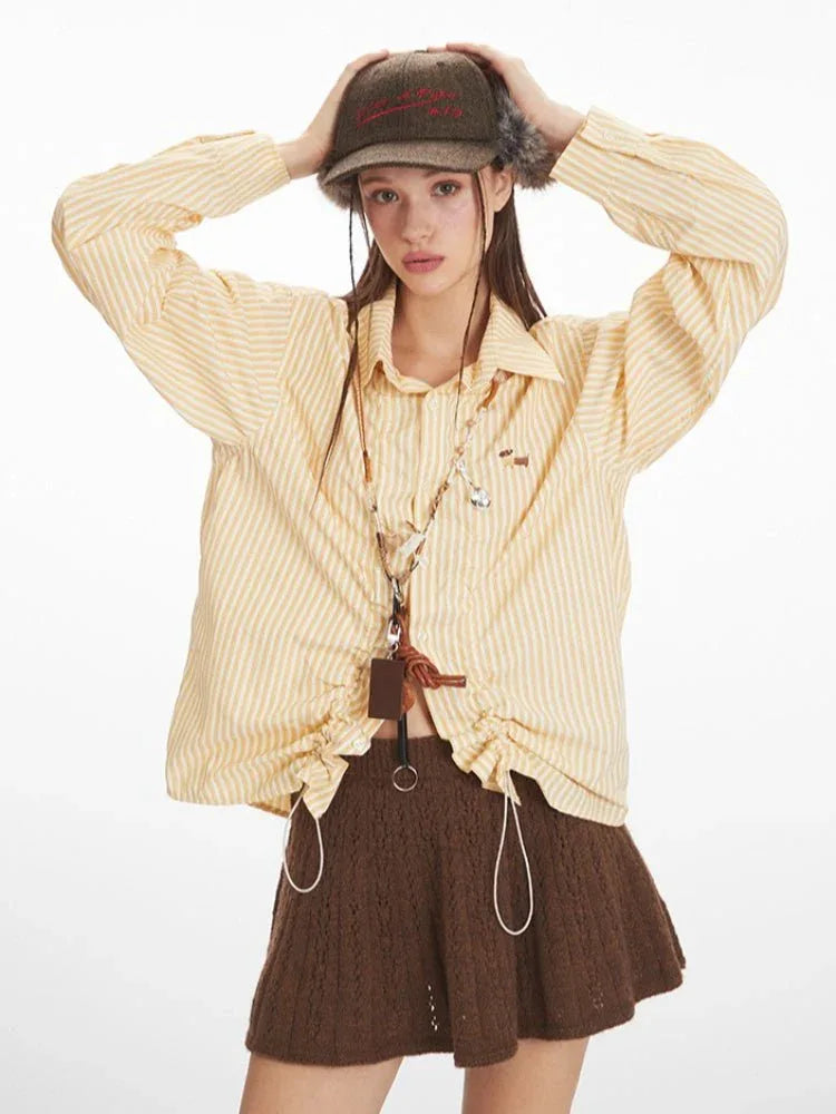 Woman wearing a yellow and white striped long-sleeved collared shirt with ruched drawstring detail and a brown knit skirt, wearing a hat and adjusting it.