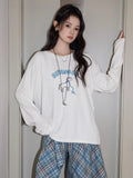 Full body shot of a woman in a white long sleeve graphic t-shirt and jeans, styled with a plaid shirt.