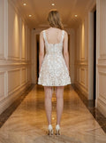 Back view of a woman in a white sleeveless floral applique short dress with a scoop neckline and a zipper closure.