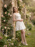 Woman leaning against a stone wall adorned with roses, wearing a white sleeveless floral applique short dress with a scoop neckline.