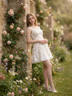 Woman leaning against a stone wall adorned with roses, wearing a white sleeveless floral applique short dress with a scoop neckline.