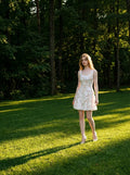 Woman walking on a grassy lawn in a forest setting, wearing a white sleeveless floral applique short dress with a scoop neckline.