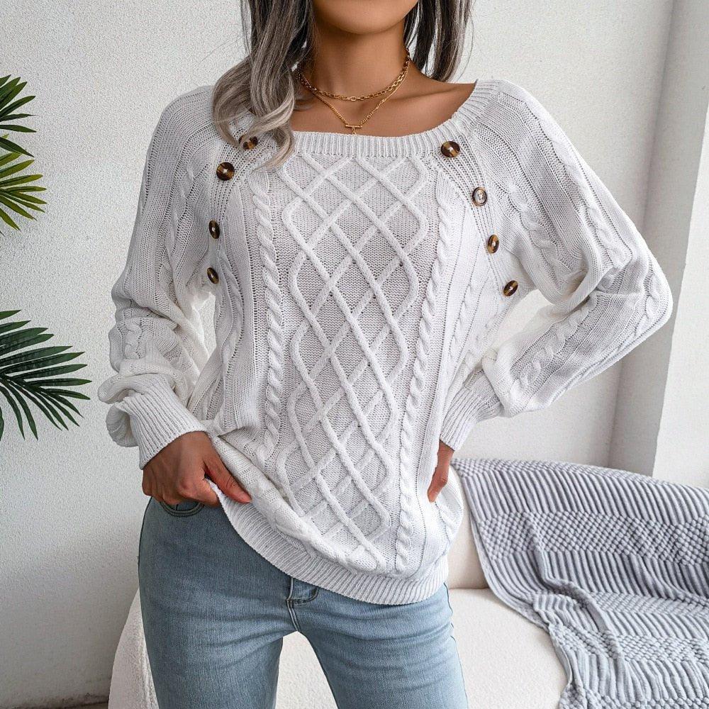 Woman wearing a white cable knit sweater with a boat neckline and decorative buttons, paired with blue jeans.